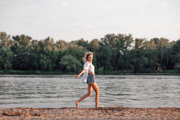 A young woman running against the background of the river. A beautiful happy blonde in a white top and shirt and denim shorts runs along the beach. 