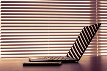 Modern workplace with laptop computer and cellphone liying on table above window with blinds. Warm sunset light with deep shadows.