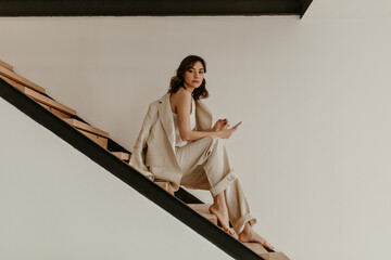 Young brunette curly woman in beige suit sits on wooden stairs. Pretty tanned lady in linen pants and jacket holds phone.