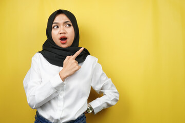 Beautiful young asian muslim woman shocked, surprised, wow expression, with hand pointing to empty space, presenting something, presenting product, isolated on yellow background