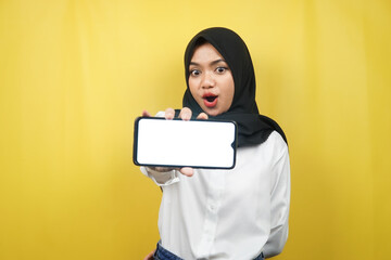 Naklejka premium Beautiful young asian muslim woman shocked, surprised, wow expression, hand holding smartphone with white or blank screen, promoting app, promoting product, presenting something, isolated