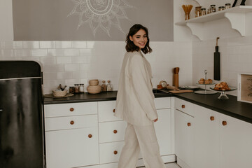 Tanned brunette lady in suit smiles widely at home. Charming woman in home linen outfit moves at kitchen in good mood.
