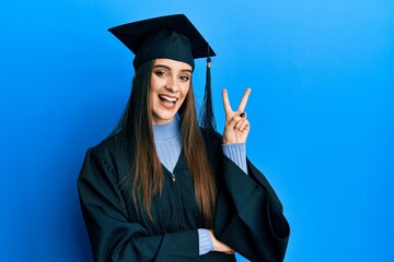 Beautiful brunette young woman wearing graduation cap and ceremony robe smiling with happy face winking at the camera doing victory sign. number two.