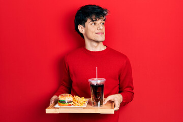 Handsome hipster young man eating a tasty classic burger with fries and soda smiling looking to the side and staring away thinking.