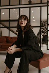 Joyful excited woman in stylish silk pants and black jacket sits on leathered brown chair at home....