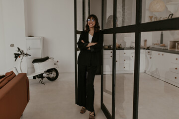 Full-length portrait of brunette curly woman in eyeglasses, stylish pants, black jacket and white top smiling sincerely and leaning on window in living room.