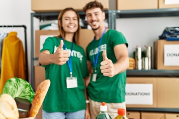 Young hispanic volunteer couple smiling happy doing ok sign with thumbs up at charity center.