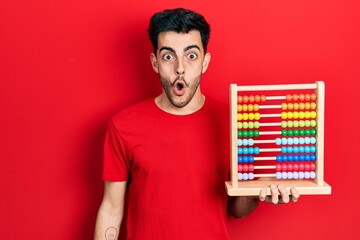 Young hispanic man holding traditional abacus scared and amazed with open mouth for surprise, disbelief face - obrazy, fototapety, plakaty