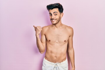 Fototapeta premium Young hispanic man wearing swimwear shirtless smiling with happy face looking and pointing to the side with thumb up.