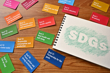 Fototapeta premium There is a card with the 17 goals of the SDGs on the desk. Beside it, there is a sketchbook with the letters of the SDGs.