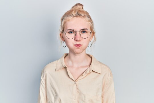 Beautiful caucasian woman with blond hair wearing casual look and glasses puffing cheeks with funny face. mouth inflated with air, crazy expression.