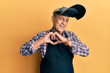 Handsome senior man with grey hair wearing welding protection mask smiling in love doing heart...