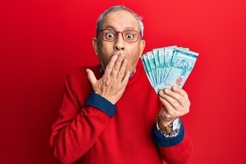 Handsome senior man with grey hair holding 50 malaysia ringgit banknotes covering mouth with hand,...
