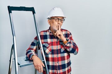Handsome mature handyman close to construction stairs wearing hardhat asking to be quiet with finger on lips. silence and secret concept.