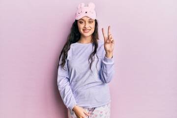 Beautiful middle eastern woman wearing sleep mask and pajama smiling with happy face winking at the camera doing victory sign. number two.