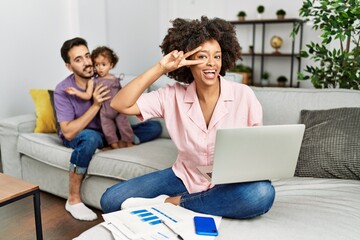 Mother of interracial family working using computer laptop at home doing peace symbol with fingers over face, smiling cheerful showing victory