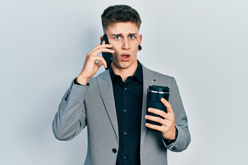 Young caucasian boy with ears dilation using smartphone and drinking a cup of coffee in shock face, looking skeptical and sarcastic, surprised with open mouth