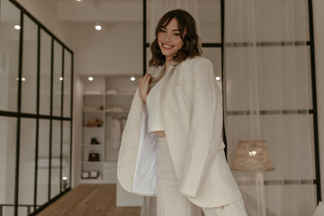 Attractive young brunette woman in stylish beige jacket and pants smiles in cozy room. Happy lady...