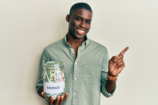 Young african american man holding jar with savings smiling happy pointing with hand and finger to the side