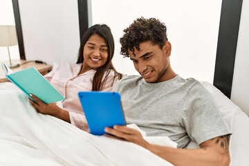 Fototapeta premium Young latin couple reading book using touchpad sitting on the bed at bedroom.