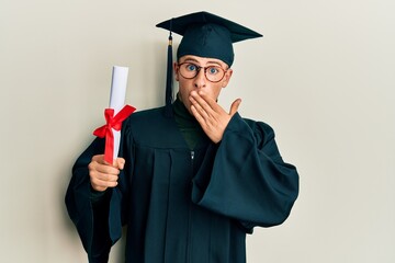 Young caucasian man wearing graduation cap and ceremony robe holding diploma covering mouth with hand, shocked and afraid for mistake. surprised expression