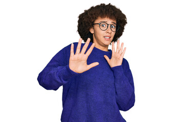 Young hispanic girl wearing casual winter sweater and glasses afraid and terrified with fear expression stop gesture with hands, shouting in shock. panic concept.