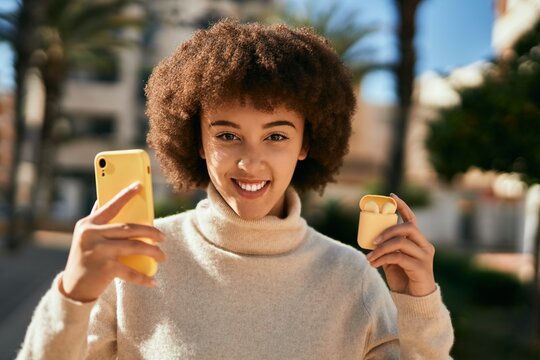 Young hispanic girl smiling happy using smartphone and holding earphones at the city.