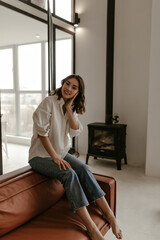 Brown-eyed brunette woman in white shirt and denim pants sits on brown leathered sofa and looks into camera with smile.