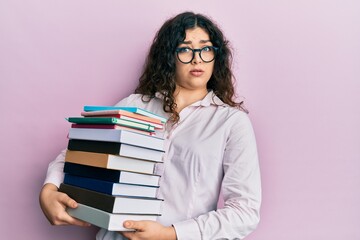 Young brunette woman with curly hair holding a pile of books clueless and confused expression. doubt concept.