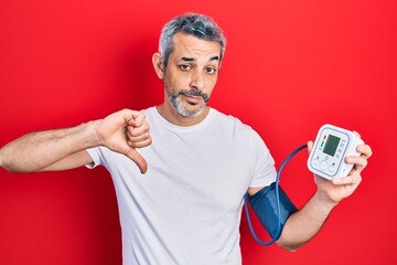 Handsome middle age man with grey hair using blood pressure monitor with angry face, negative sign...