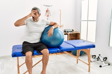 Middle age hispanic man at pain recovery clinic holding pilates ball peeking in shock covering face and eyes with hand, looking through fingers with embarrassed expression.