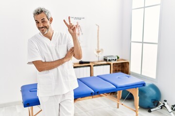 Middle age hispanic therapist man working at pain recovery clinic smiling looking to the camera showing fingers doing victory sign. number two.