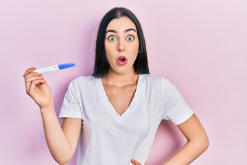 Beautiful woman with blue eyes holding pregnancy test result scared and amazed with open mouth for surprise, disbelief face