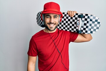 Young hispanic man using headphones holding skate looking positive and happy standing and smiling with a confident smile showing teeth