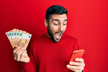 Young hispanic man using smartphone holding 50 euros banknotes celebrating crazy and amazed for...