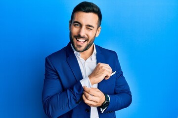 Young hispanic businessman wearing business jacket putting on shirt button winking looking at the...