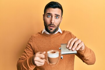 Young hispanic man drinking a cup of coffee with alcohol in shock face, looking skeptical and...