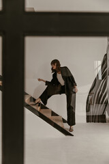 Woman in black suit stands on wooden stairs in apartment. Brunette curly lady in stylish pants and jacket poses in white room.