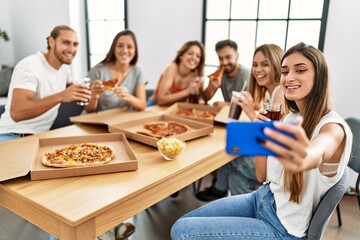Group of young people smiling happy eating italian pizza and make selfie by the smartphone at home