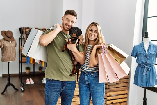 Young hispanic customer couple smiling happy holding shopping bags and dog at clothing store.