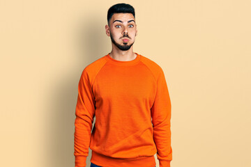 Young hispanic man with beard wearing casual orange sweater puffing cheeks with funny face. mouth inflated with air, crazy expression.