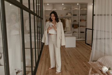 Elegant lady in linen beige pants and oversized jacket holds phone and reads message. Pretty curly lady in suit stands in her dressing room.