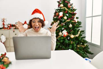 Middle age hispanic woman using laptop sitting by christmas tree celebrating victory with happy smile and winner expression with raised hands