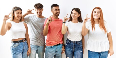 Group of young friends standing together over isolated background smiling and confident gesturing with hand doing small size sign with fingers looking and the camera. measure concept.