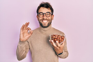 Handsome man with beard holding bowl of chestnuts doing ok sign with fingers, smiling friendly gesturing excellent symbol