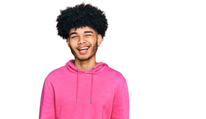Young african american man with afro hair wearing casual pink sweatshirt winking looking at the...