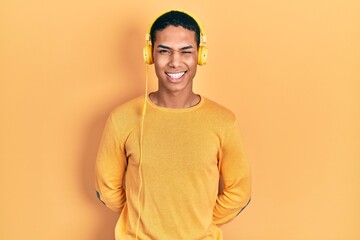 Young african american guy listening to music using headphones winking looking at the camera with...