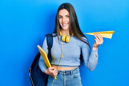 Young brunette teenager student holding books and paper plane smiling with a happy and cool smile on face. showing teeth.