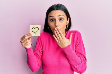 Young hispanic woman holding heart reminder covering mouth with hand, shocked and afraid for...
