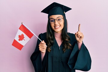 Young hispanic woman wearing graduation uniform holding canada flag smiling with an idea or question pointing finger with happy face, number one